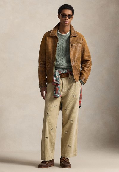 Giacca di pelle marrone, maglione verde a trecce, pantaloni beige di velluto a coste con dettagli ricamati, camicia a quadri annodata in vita, scarpe marroni.