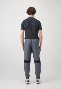 Schwarzes Kurzarm-Sportshirt kombiniert mit grauen Jogginghosen, die schwarze Streifen und elastische Bündchen haben. Weiße Sportschuhe runden das Outfit ab.