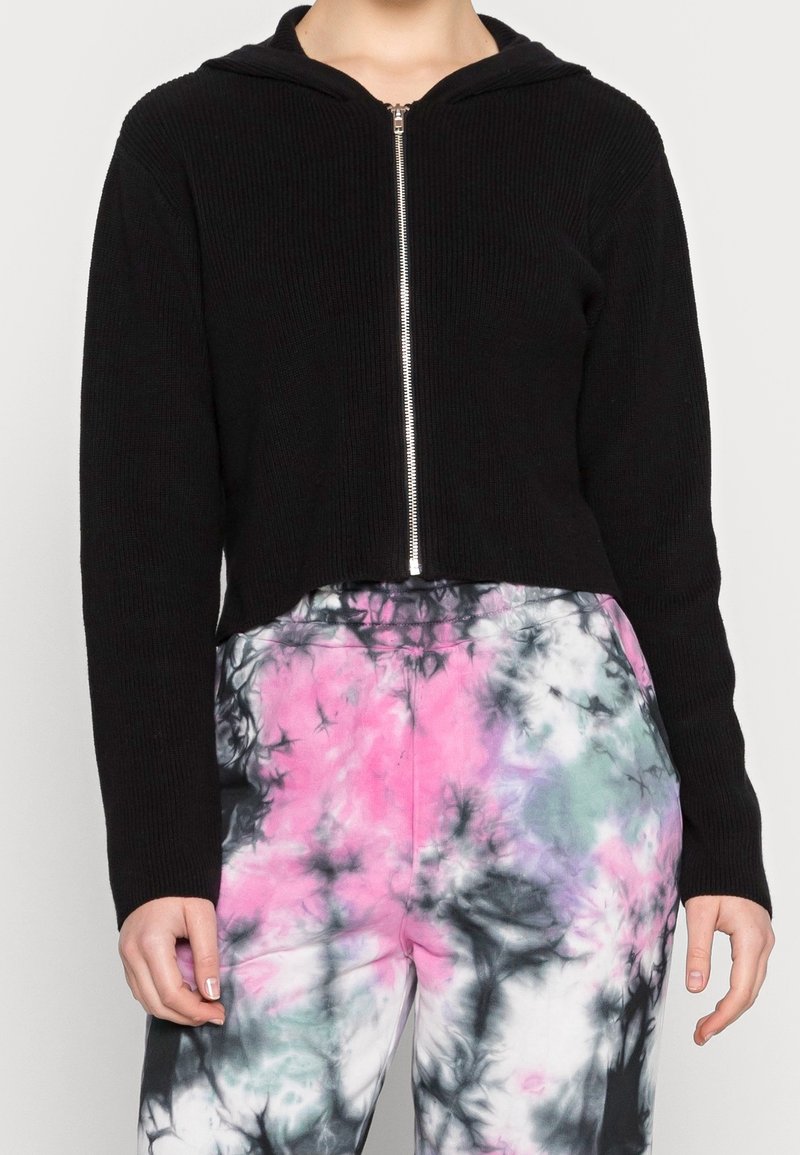 Sweat à capuche noir court avec fermeture éclair et texture côtelée, accompagné d'un pantalon tie-dye multicolore aux motifs rose, noir et gris.