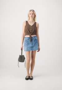 Brązowy prążkowany crop top z guzikami, dżinsowa mini spódnica z postrzępionym dołem, czarne baleriny z ćwiekami i wzorzysty worek na ramię. Modelka stoi na tle białym.