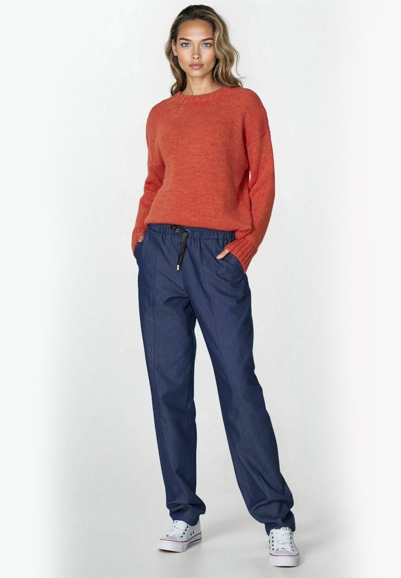 Kobieta stoi z rękami w kieszeniach, ubrana w sweter w kolorze rdzawym, luźne niebieskie spodnie z gumką w pasie oraz białe sneakersy na jednolitym tle.