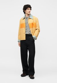 Άνδρας φορά κίτρινο και πορτοκαλί μπουφάν πάνω από καρό πουκάμισο με μαύρο παντελόνι φαρδιάς γραμμής και σκούρα παπούτσια, στέκεται και κοιτάζει δεξιά.