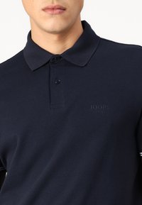 Temno modra polo majica z teksturirano tkanino, z gumbom z zloženo plošče in subtilnim embosiranim logotipom na prsih. Kratke rokave.