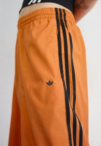 Oranži sporta šorti, izgatavoti no gludas auduma, ar melnām svītrām pa sāniem un mazu melnu logotipu kreisajā kājā.
