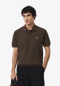 Polo shirt marrone in morbido cotone con colletto, due bottoni e logo verde Lacoste sul lato sinistro del petto. Indossato con pantaloni neri.
