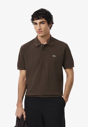 Brązowa koszulka polo wykonana z miękkiej bawełny, z kołnierzykiem, dwoma guzikami i zielonym logo Lacoste na lewej piersi. Noszona z czarnymi spodniami.