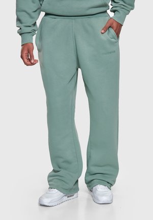 Mees, kes kannab salvei rohelisi dressipükse ja sobivat dressipluusi, valged vabaajajalatsid, seisab ühe käega taskus ühevärvilise tausta ees.