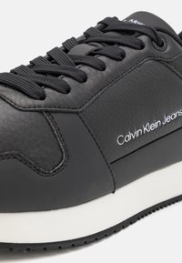 Svart lädersneaker med texturerad yta, platta svarta snörning, vit sula och subtil logotyp på sidan. Slät, modern design.