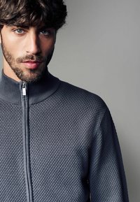 Grå zip-up tröja i texturerad tyg, hög krage och en slät dragkedja. Tight passform. Subtil geometrisk mönstring genom hela tröjan.