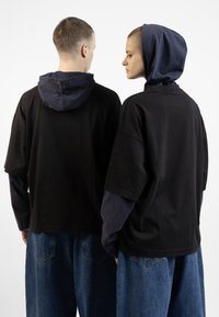 Tricouri negre supradimensionate cu accente de capuci bleu marin, țesătură texturată și stil lejer, asortate cu pantaloni din blug care au buzunare pe spate.