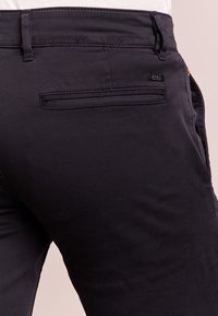 Pantalon en tissu noir à texture lisse, avec une poche arrière et une petite étiquette logo, présenté de la taille jusqu'en bas.