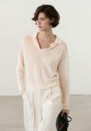 Jeune femme aux cheveux courts portant un pull pêche clair et un pantalon crème, tenant un petit sac à main noir tressé, regardant vers la gauche.