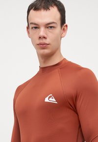 Rash guard de manga larga marrón hecho de material liso y elástico que presenta un logo blanco en el pecho, con costuras planas y un diseño ajustado.