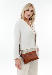 Femme blonde en blouse blanche et pantalon, portant un sac bandoulière en cuir marron avec poche zippée à l'avant, posant devant un fond blanc.