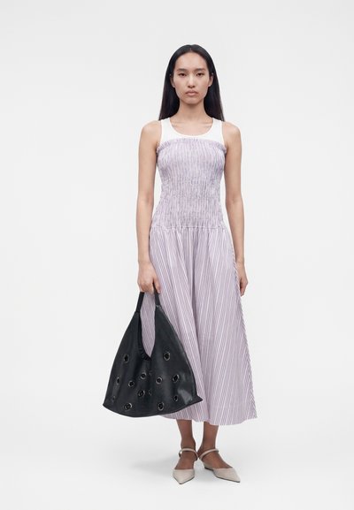 Γυναίκα στέκεται φορώντας μωβ ριγέ φόρεμα χωρίς μανίκια και κρατώντας μια μεγάλη μαύρη τσάντα με κυκλικά κοψίματα.