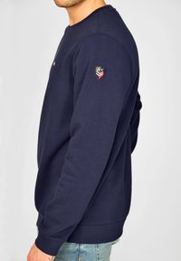 Granatowy sweatshirt wykonany z bawełny, z okrągłym dekoltem, ściągaczami przy rękawach oraz metką z logo na lewym rękawie. Prosty i klasyczny design.