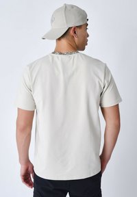 Camiseta de manga corta color beige claro con una textura suave, que presenta un escote estampado y un corte relajado. Se lleva con una gorra gris.