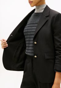 Černý blazer se zlatými knoflíky, pod kterým se nachází texturovaný tmavě šedý pruhovaný svetr. Interiér má hladkou černou podšívku s jemným vzorem.