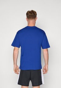 Blå bomulds t-shirt med rund halsudskæring, korte ærmer og lige kant, set fra bagsiden. Bæret med sorte shorts med hvide accenter.