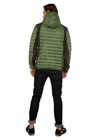 Giacca puffer verde con cappuccio e pannelli laterali grigi, con una texture trapuntata, indossata sopra jeans scuri e sneakers nere.