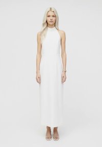 Hosszú szőke hajú nő, ujjatlan, magas nyakú fehér maxiruha és áttetsző, nyitott orrú magassarkú cipő visel, egy sima fehér háttér előtt áll.