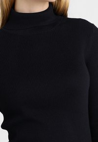 Pull en col roulé noir à côtes, fabriqué dans un tissu lisse et extensible. Présente un design ajusté et un col haut pour plus de chaleur et de couverture.