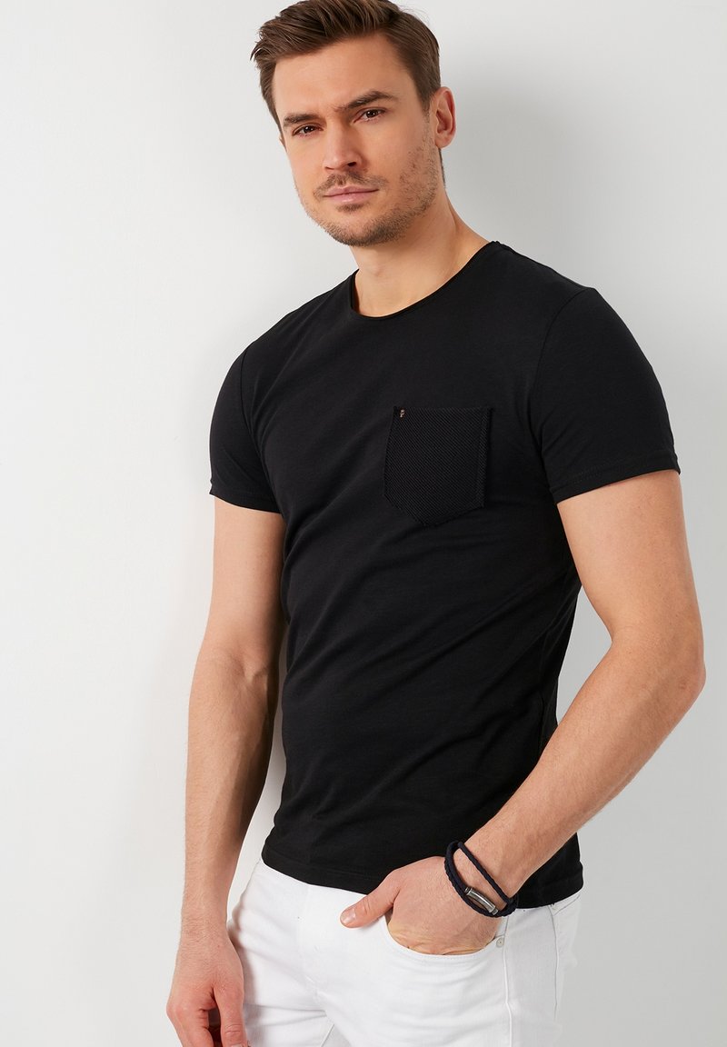 T-shirt nera a maniche corte con dettagli a tasca testurizzati. Vestibilità standard con scollatura rotonda. Indossata con pantaloni bianchi e un bracciale scuro.