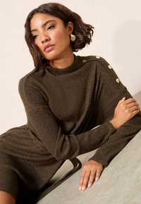 Abito a maglia a coste verde scuro, con maniche lunghe, colletto alto e dettagli con bottoni sulle spalle, rifinito con un effetto cintura annodata.