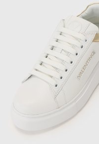 Hvit lær sneakers med flate, hvite skolisser, teksturert gummisåle, gullaksent på hælen og preget logo detaljer på siden.