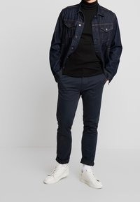 Homme portant une veste en denim foncé, un col roulé noir, un pantalon bleu marine, des chaussettes blanches et des baskets blanches, se tenant contre un fond uni.
