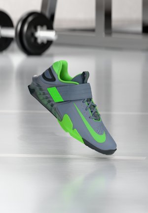 Grauer Sportschuh mit einem grünen Akzentriemen und einer Sohle, ausgestattet mit einem strukturierten Obermaterial und perforierten Details für Atmungsaktivität.