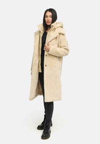 Cappotto lungo beige con texture in pelliccia, con cappuccio, due bottoni e tasche laterali, indossato sopra una giacca leggera con zip e jeans neri.