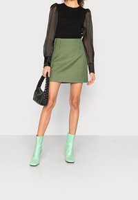 Jeune femme se tenant debout, portant un haut noir à manches transparentes, une jupe à carreaux verts et des bottines vert menthe, tenant un sac à main noir.