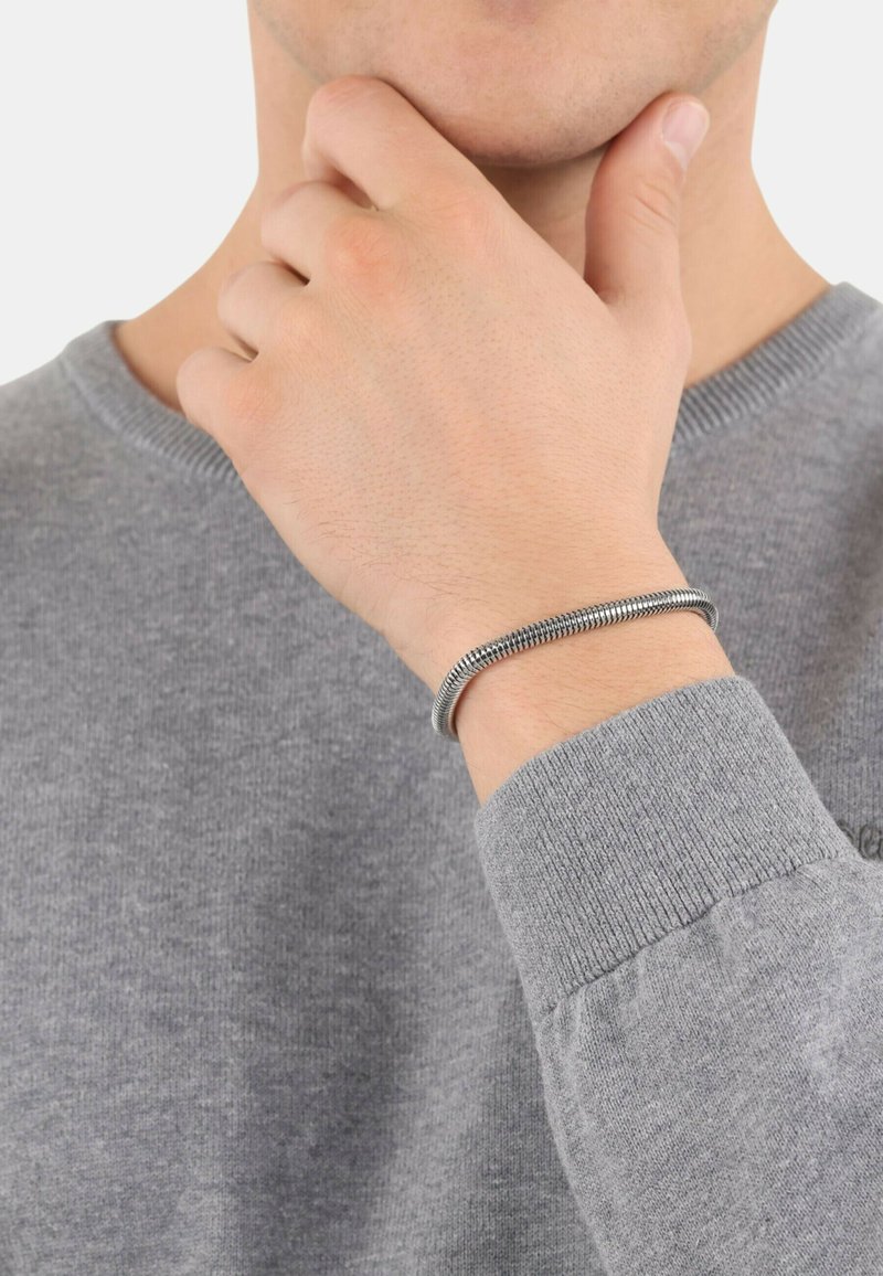 Pulsera de metal plateado con un diseño cilíndrico y suave, llevada en una muñeca. El portador viste un suéter gris con puños acanalados.
