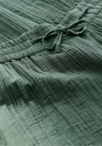 Tessuto verde con una texture stropicciata e linee sottili. Presenta una vita arricciata con un dettaglio a fiocco. Materiale leggero e morbido.