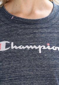 Primo piano di una persona che indossa una felpa Champion grigio scuro con il logo bianco e rosa sul petto.