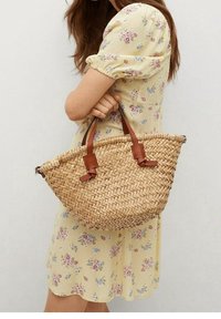 Bolso de paja tejido con acentos de cuero marrón, forma redondeada y asas dobles. Muestra un vestido de estampado floral al fondo.
