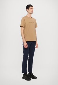 Brown cotton T-shirt with embossed brand logo, paired with navy trousers and black athletic shoes. Model stands against a white background.
