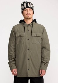 Veste chemise en velours côtelé vert avec deux poches poitrine, boutons noirs, capuche réglable et texture côtelée, portée avec un bonnet à motifs.
