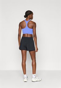 Blå atletisk sports-bh med racerback-design, kombineret med sorte shorts. Modellen vender ryggen til, hvilket fremhæver stoffet og pasformen.