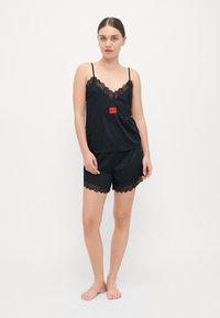 Musta satiininen yöasu, jossa on pitsiyksityiskohtia. Camisole-yläosassa on V-pääntie ja säädettävät olkaimet; shortseissa on pitsireuna helman kohdalla. Punainen logoaksentti.