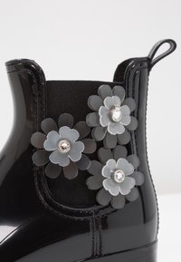 Bottine noire brillante avec un panneau latéral élastique décoré de trois embellissements floraux translucides, chacun orné d'un strass central.