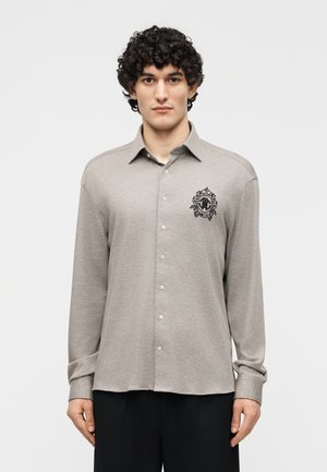 Άνδρας με σγουρά μαλλιά που φοράει ανοιχτό γκρι πουκάμισο με μακριά μανίκια και μαύρο κεντημένο εμβλημα στον αριστερό θώρακα.