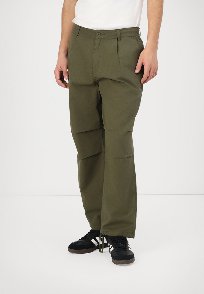 Calças cargo verde-oliva com corte relaxado, apresentando pregas frontais e bolsos laterais, combinadas com ténis atléticos pretos.