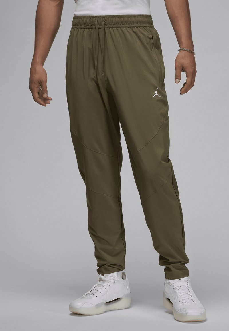 Mand iført olivengrønne træningsbukser med Jordan-logo og hvide sneakers, stående mod en ensfarvet grå baggrund.
