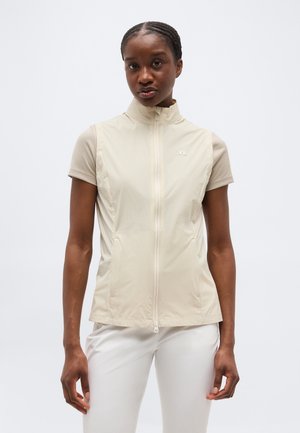 Γυναίκα με κοτσίδες, που φοράει μπεζ αμάνικο γιλέκο με φερμουάρ πάνω από κοντομάνικο πουκάμισο και λευκό παντελόνι, στέκεται μπροστά σε απλό φόντο.