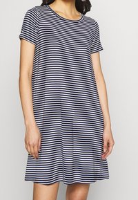 Kobieta ubrana w sukienkę z krótkim rękawem w poziome niebieskie i białe paski, z okrągłym dekoltem, stojąca na tle jasnego, jednolitego tła.