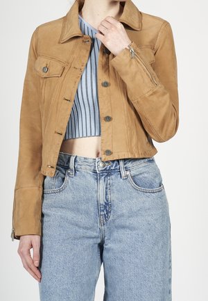 Mujer con chaqueta corta de ante color tostado sobre top corto de rayas azules y grises con vaqueros de tiro alto azul claro, una mano cerca del cuello.