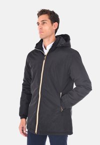 Uomo che indossa una giacca invernale nera con cappuccio e una cerniera a strisce multicolori, con le mani in tasca, che guarda di lato su uno sfondo bianco.