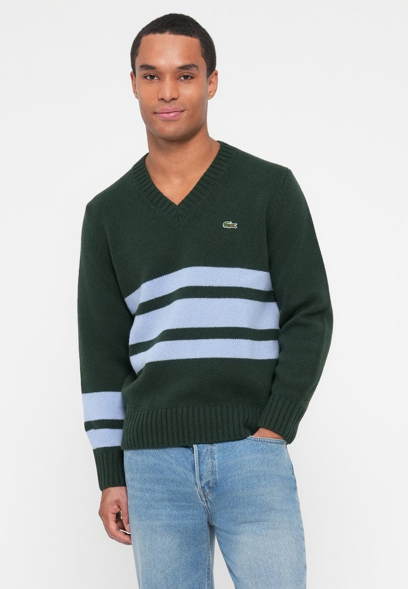 Grøn strikket sweater med v-udskæring, med horisontale lyseblå og mørkegrønne striber samt et lille logo på brystet. Ribbet manchetter og kant.